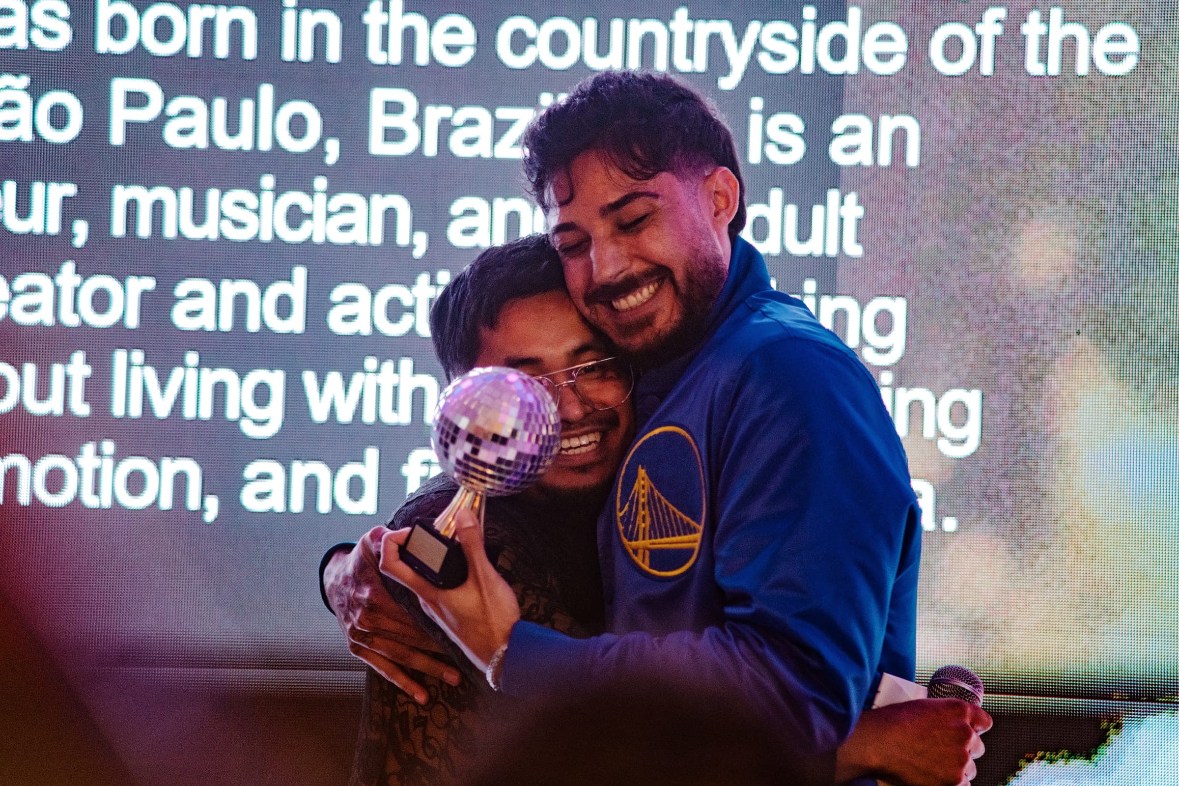 Two friends embracing on stage, one holding a disco-ball award